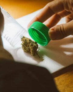 Hand opening a clear plastic container with a green lid, revealing a cannabis bud on a white surface.